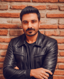 a man wearing a black leather jacket with his arms crossed stands in front of a brick wall