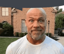 a bald man with a beard and a white shirt is making a funny face in front of a brick house .