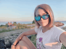 a woman wearing sunglasses and a t-shirt with a unicorn on it sits in front of a body of water