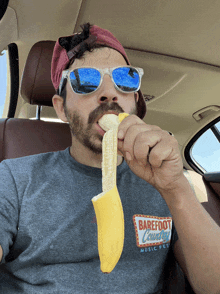 a man in a car wearing a shirt that says barefoot country is eating a banana