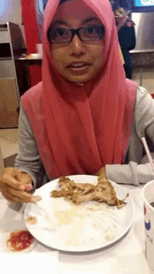 a woman wearing glasses and a pink hijab is sitting at a table with a plate of food in front of her