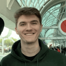 a young man wearing a green hoodie is smiling in front of a glass building .