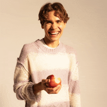 a young man wearing a striped sweater is holding an apple and smiling