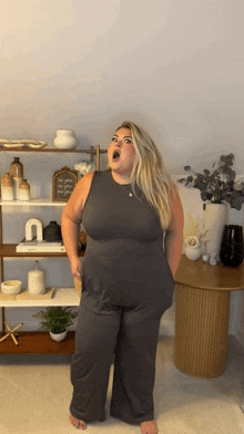 a woman in a grey jumpsuit is standing in front of a shelf with vases on it .