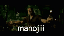 a man is standing in a dark room with his arms outstretched and the word manojiii written in white letters .