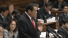 a man in a suit and red tie is giving a speech in front of a microphone in a parliament .