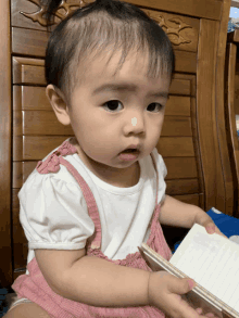 a baby girl in a pink and white dress is holding a book