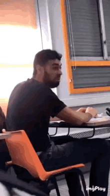 a man with a beard is sitting at a desk in a classroom with a window in the background .