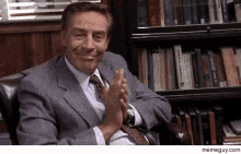 a man in a suit and tie is clapping his hands in front of a bookshelf filled with books .