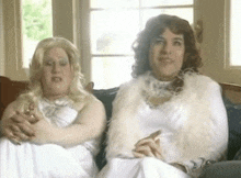 two women are sitting on a couch and one is wearing a white dress