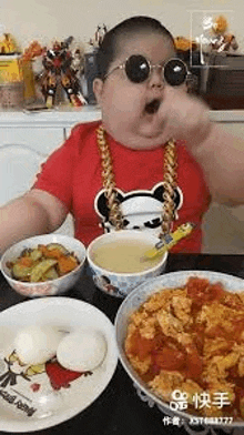 a baby wearing sunglasses and a gold chain is eating food from a bowl .