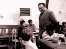 a man in a suit and tie is standing in front of a group of children sitting at tables in a classroom .