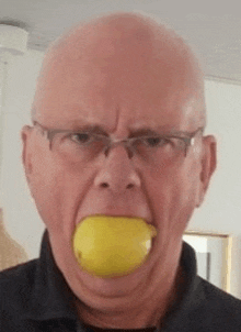 a bald man with glasses is biting a lemon in his mouth .