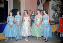 a group of women in blue and green dresses are standing in a room with a man in a suit behind them