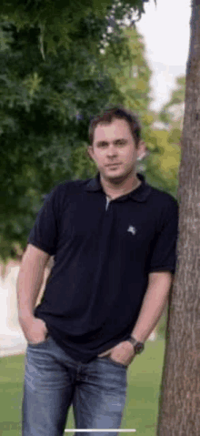 a man in a black polo shirt is leaning against a tree .