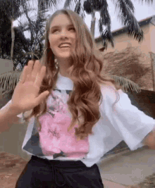 a woman with long hair is wearing a white t-shirt with a pink flower on it and waving at someone .