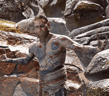 a man with a beard and tattoos on his torso is standing in front of a pile of rocks with his arms outstretched