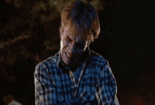 a man in a plaid shirt is sitting at a campfire at night and smiling .