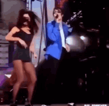 a man in a blue jacket is singing into a microphone while a woman in a black dress is dancing behind him .