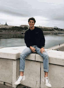 a man wearing a black sweatshirt that says ' calvin klein ' on it sits on a ledge overlooking a river