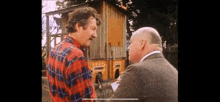 a man in a plaid shirt is talking to another man in a suit in front of a wooden building .