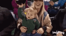 a woman is holding a baby in her lap at a baseball game and the baby is raising his hand in the air .