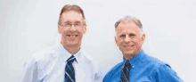 two men in blue shirts and ties are standing next to each other and smiling for the camera .