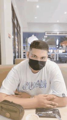 a man wearing a mask and a t-shirt that says kalinago sits at a table with his hands folded