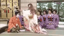 a group of women in kimonos are sitting on the floor in a room with a man in a kimono .