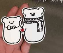 a person is holding two bear ornaments in their hand . one bear is wearing a scarf and holding a heart .