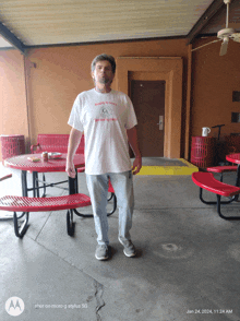 a man wearing a white t-shirt that says " simply to serve " stands in front of red picnic tables