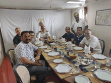 a group of men are sitting at a long table with plates of food in front of a wall with a flag that says " india "