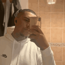 a man in a white nike hoodie is taking a selfie with his cell phone in a bathroom .