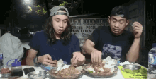 two men are sitting at a table eating food in front of a sign that says ' nasi uduk '