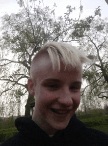 a boy with a shaved head smiles in front of a tree