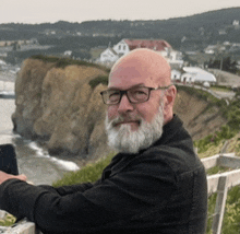 a man with glasses and a beard is sitting on a ledge overlooking a body of water