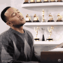 a man is singing into a microphone while playing a piano in front of a shelf full of grammy trophies .