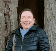 a woman wearing a black jacket and a blue scarf smiles in front of a tree