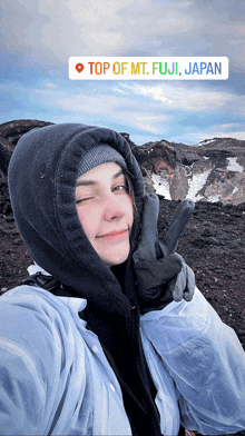 a woman wearing a hooded jacket and gloves is taking a selfie in front of top of mt. fuji japan