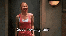 a woman in a red tank top is standing in front of a door and saying good morning , slut .