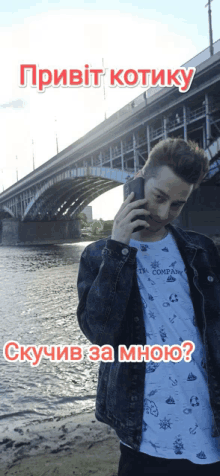 a man in a denim jacket is talking on a cell phone in front of a river