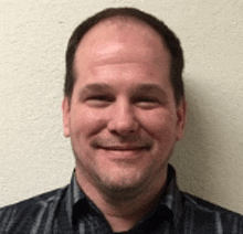 a man in a plaid shirt is smiling for the camera while standing in front of a white wall .