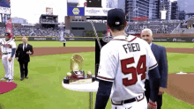 a baseball player with the number 54 on his jersey is standing on a baseball field with a trophy on a table .