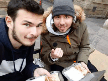 two men are sitting next to each other on a bench eating food from plastic containers .