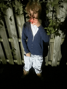 a young man in a blue hoodie is standing in front of a wooden fence smoking a cigarette