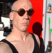 a bald man wearing sunglasses and a black tank top stands in front of a red door