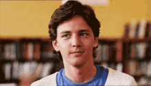 a young man in a blue shirt and white sweater is standing in front of a bookshelf in a library .