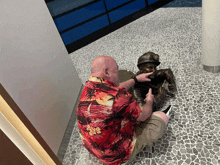 a man in a hawaiian shirt sits on the floor pointing at a statue