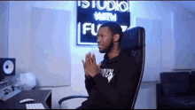 a man is sitting in a chair in front of a computer in a studio with his hands folded in prayer .