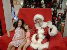 a man in a santa suit sits in a chair with two girls